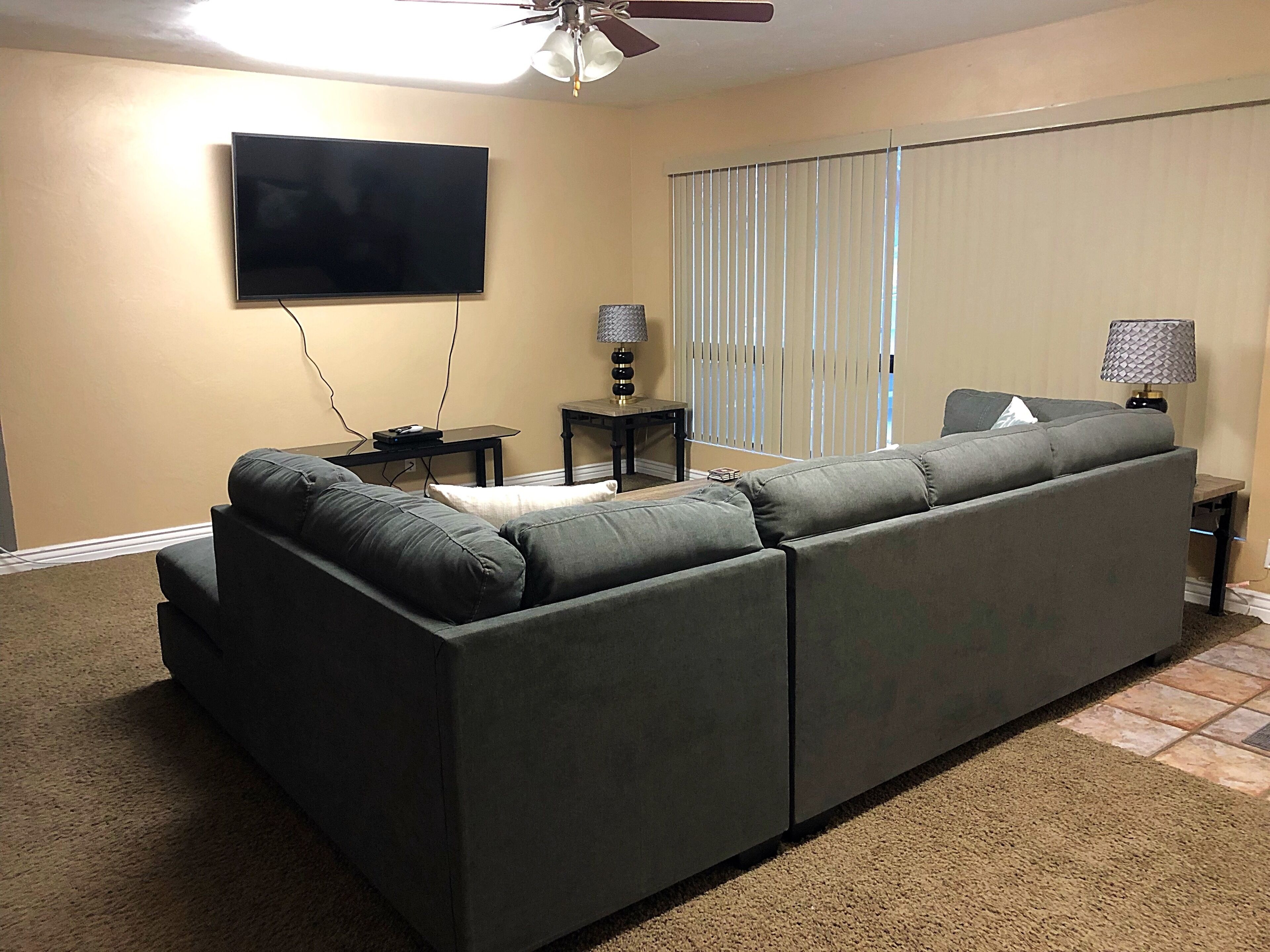 A living room with a sectional sofa, a flat-screen TV mounted on the wall, two side tables with lamps, and a ceiling fan.