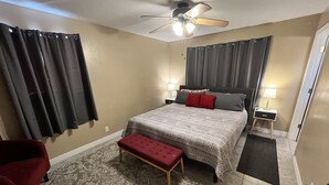 Une chambre à coucher avec un lit, une chaise rouge, un banc, une table de chevet, une lampe et un ventilateur de plafond.