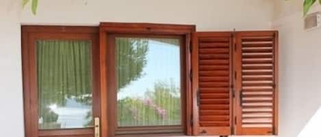 A covered outdoor seating area with a table and chairs, a wooden door with shutters, and a window with a view of greenery.