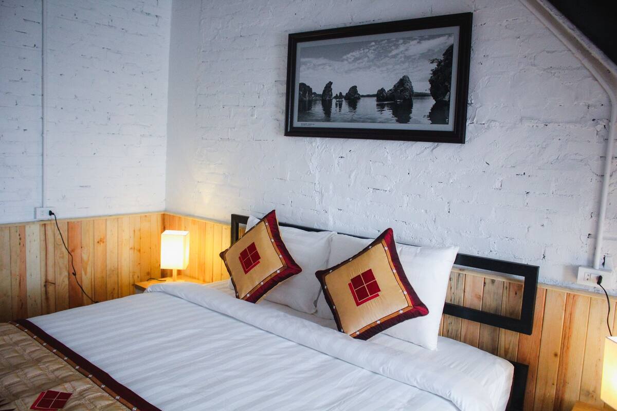 Une chambre à coucher avec une tête de lit en bois, un lit recouvert d’une literie blanche, deux coussins décoratifs, une table de chevet avec une lampe et un tableau encadré accroché au mur.