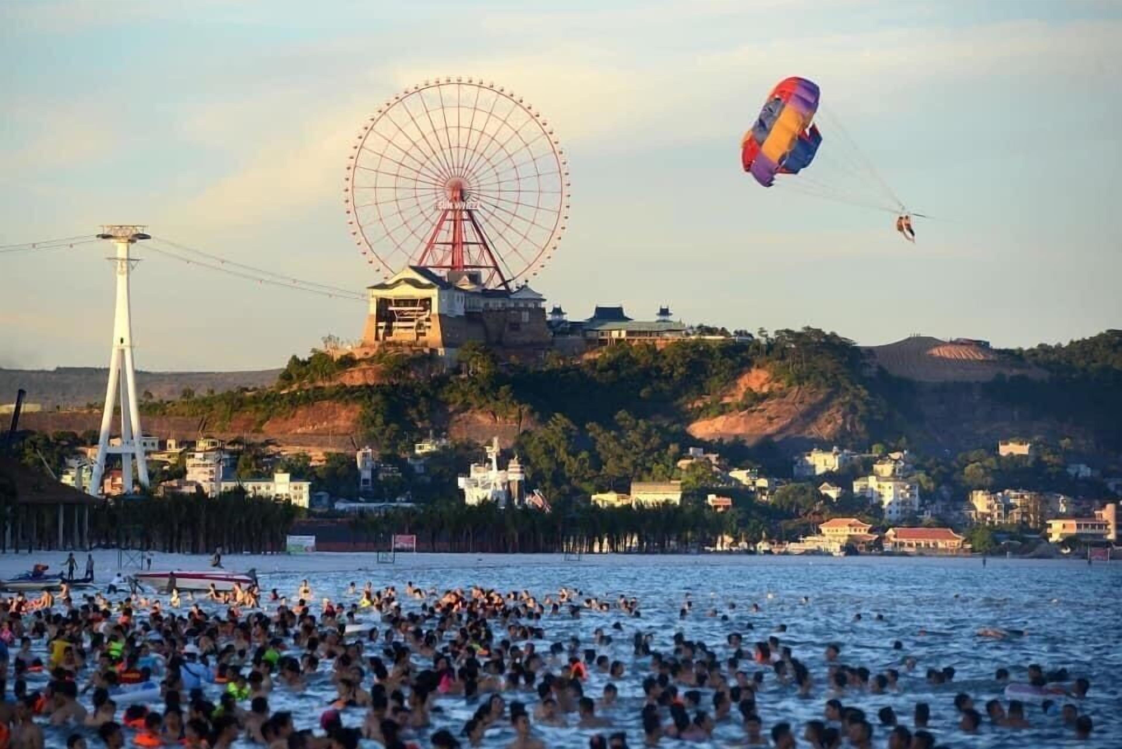 Uma praia lotada com uma grande roda-gigante e um parapente ao fundo.
