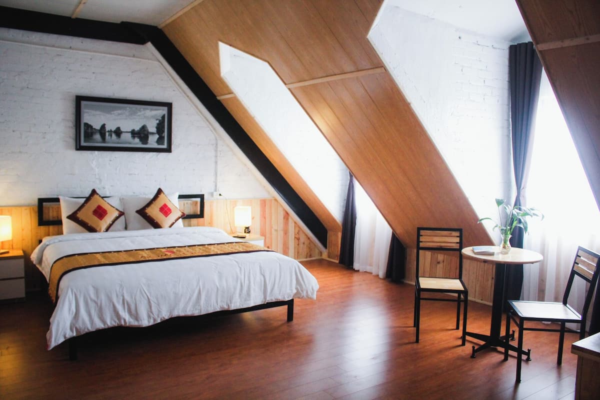 Une chambre avec un grand lit, deux tables de chevet avec des lampes, une petite table ronde avec une chaise, et une grande fenêtre avec des rideaux.