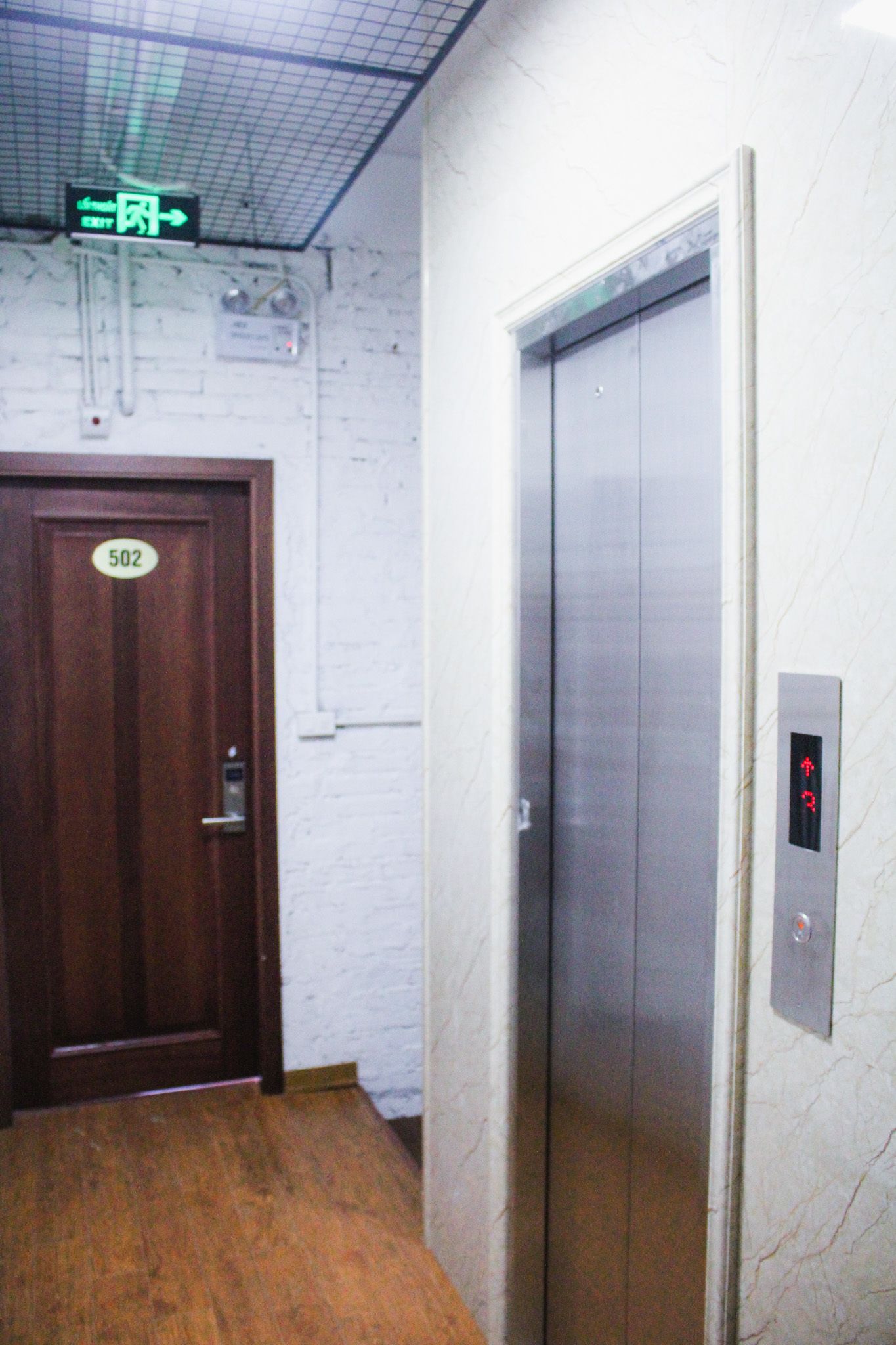 Un ascensor con pantalla digital, una puerta de madera con el número 502, y un letrero de salida verde en la parte superior.