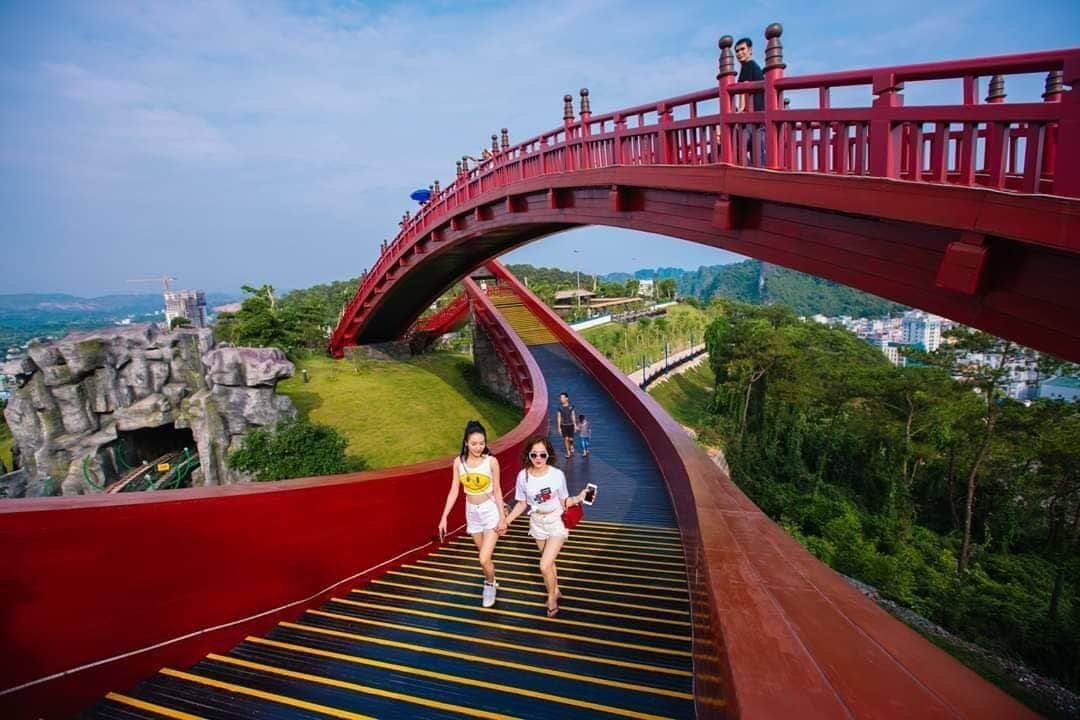 Un puente rojo de diseño curvo, un camino con líneas amarillas y una vista panorámica de la ciudad al fondo.