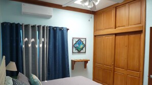 Een slaapkamer met een bed, gordijnen, een houten garderobe, een wandairconditioner en een klein plankje met een decoratief object.