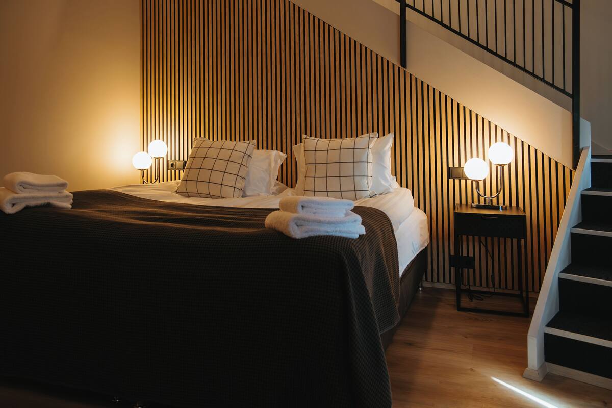 A neatly made bed with a dark bedspread and white linens, a wooden slatted headboard, and a small bedside table with a lamp.
