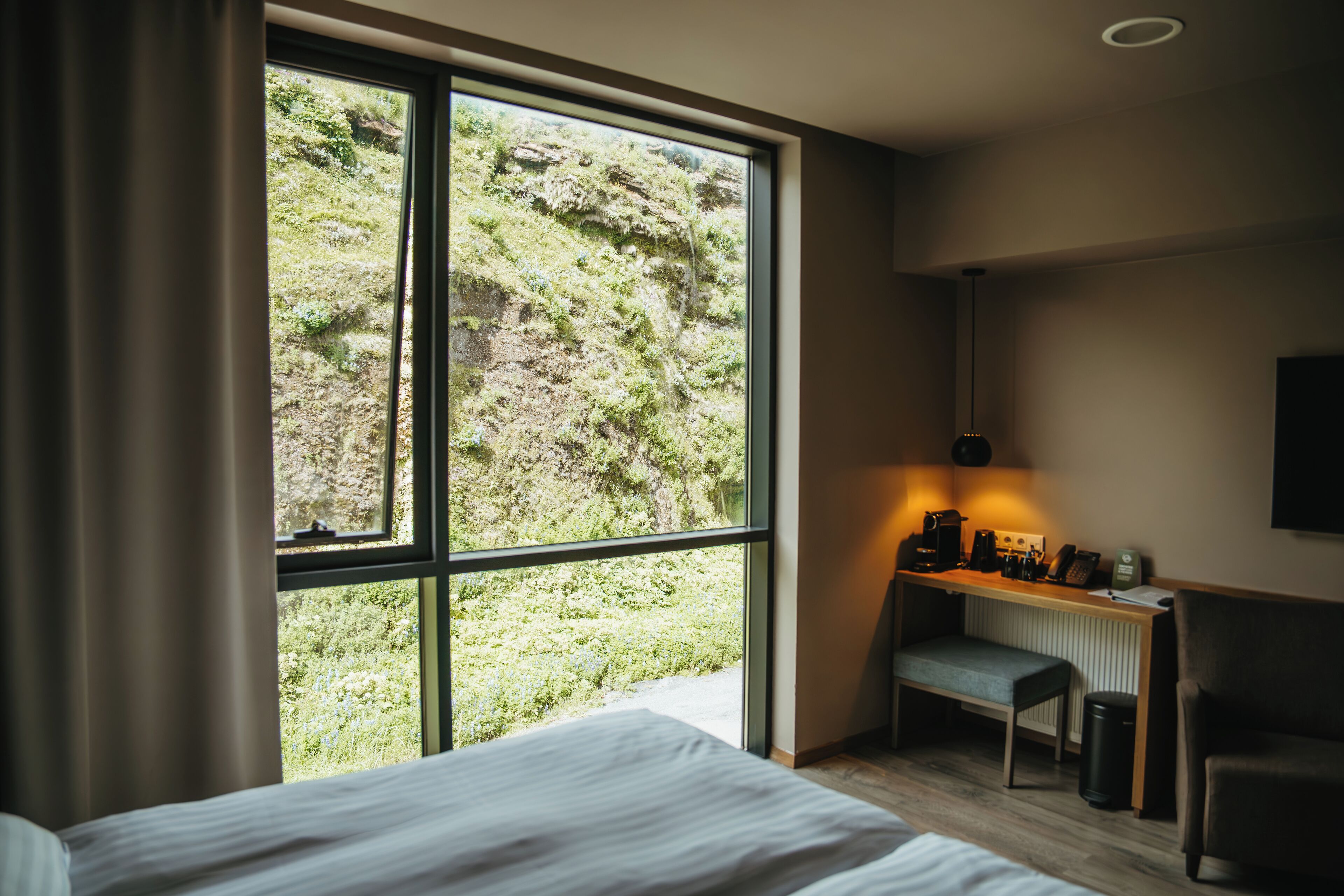 A hotel room with a large window, a bed, a desk with a chair, and a radiator.