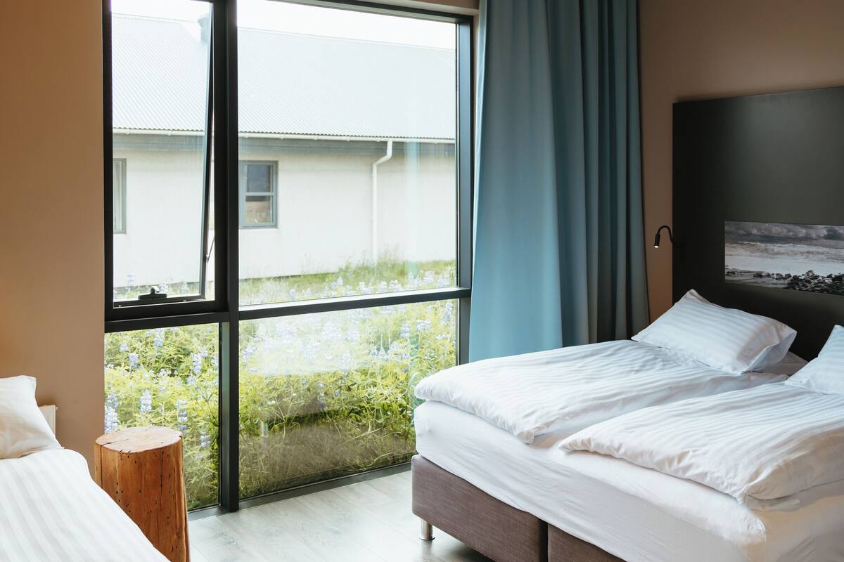 A modern hotel room with a large window, a bed, a bedside table, and a headboard.