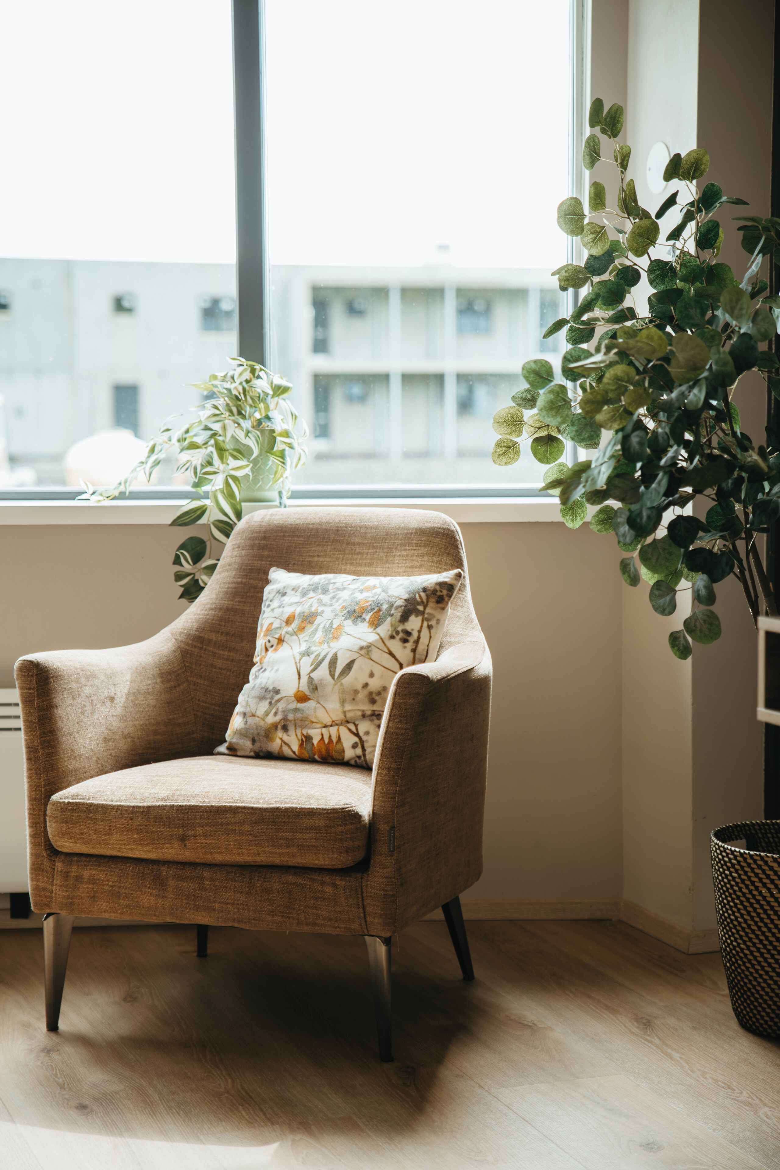 A corner room with a beige armchair, a window with a view of buildings, and a potted plant.