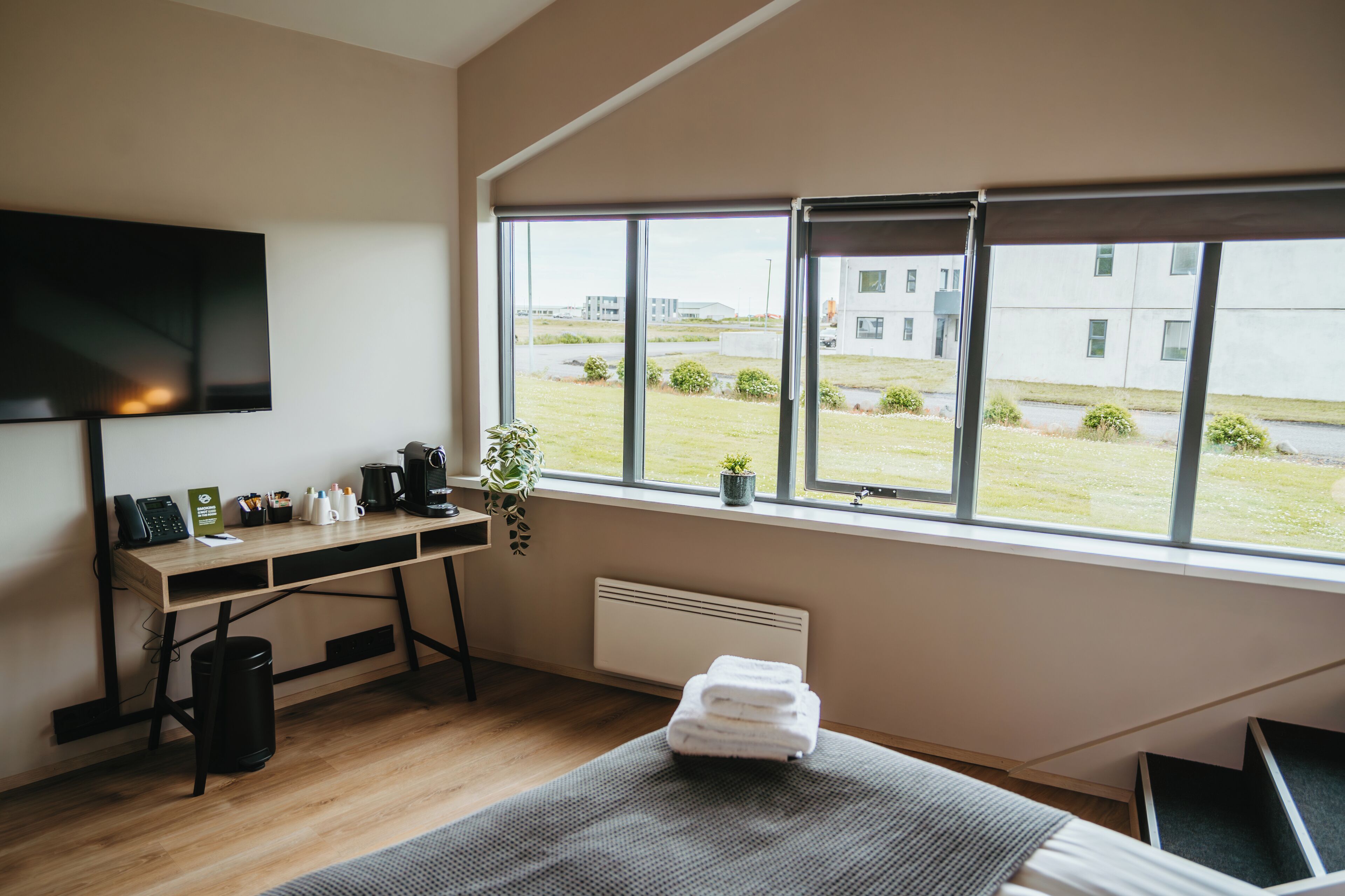 Habitación de hotel con un ventanal grande, un escritorio con televisión de pantalla plana, un radiador y una cama con manta gris.