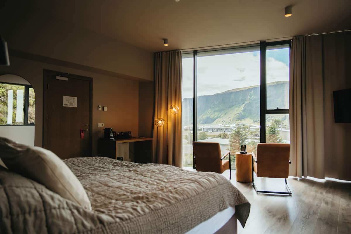 A hotel room with a large bed, a desk with a computer, a chair, and a view of mountains through the window.