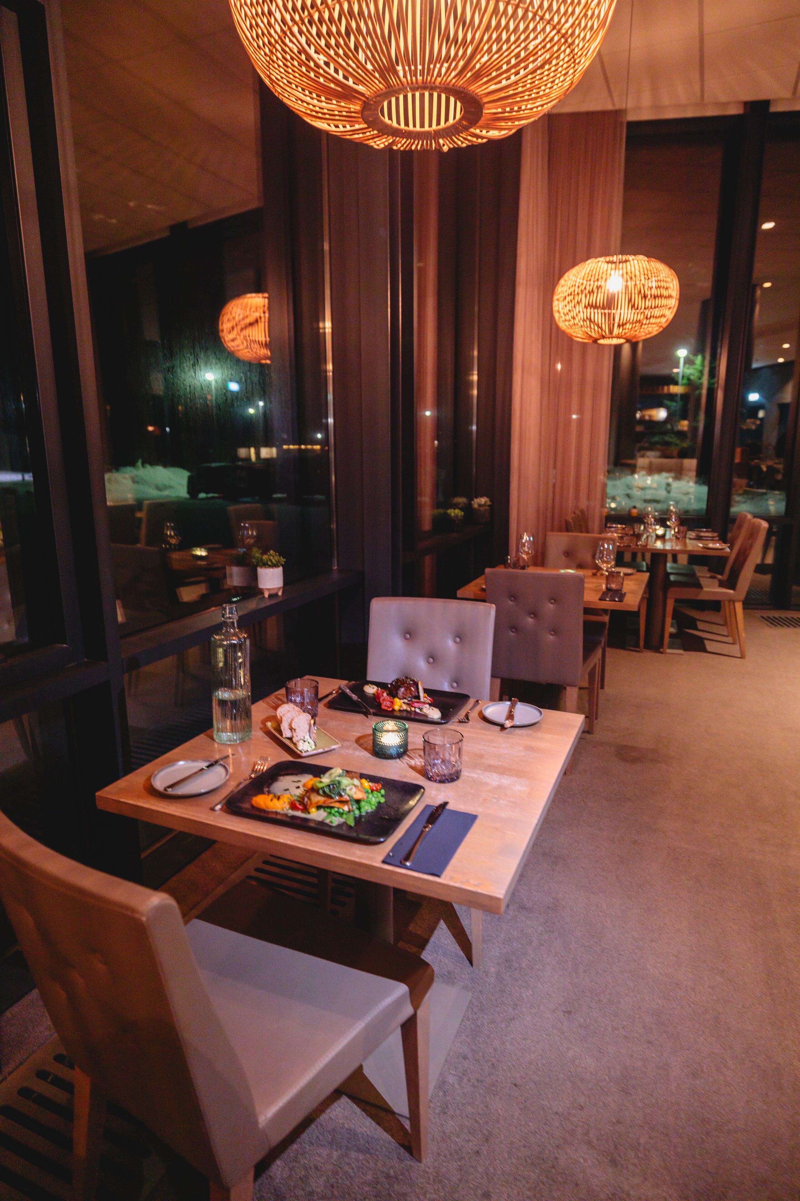 A restaurant with a long table set for dining, large windows, and hanging light fixtures.