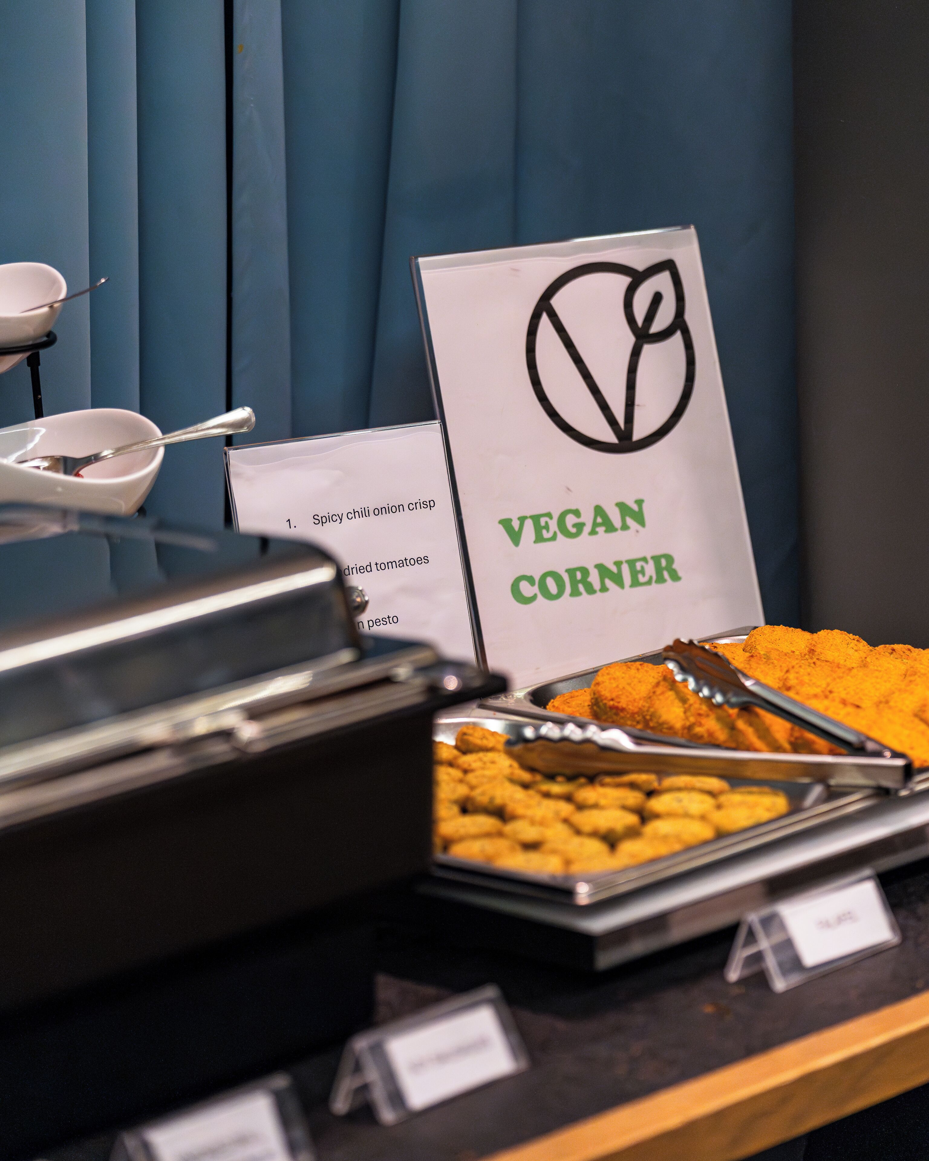 A buffet with a sign labeled "VEGAN CORNER" featuring items like spicy chili onion crisp, dried tomatoes, and pesto.