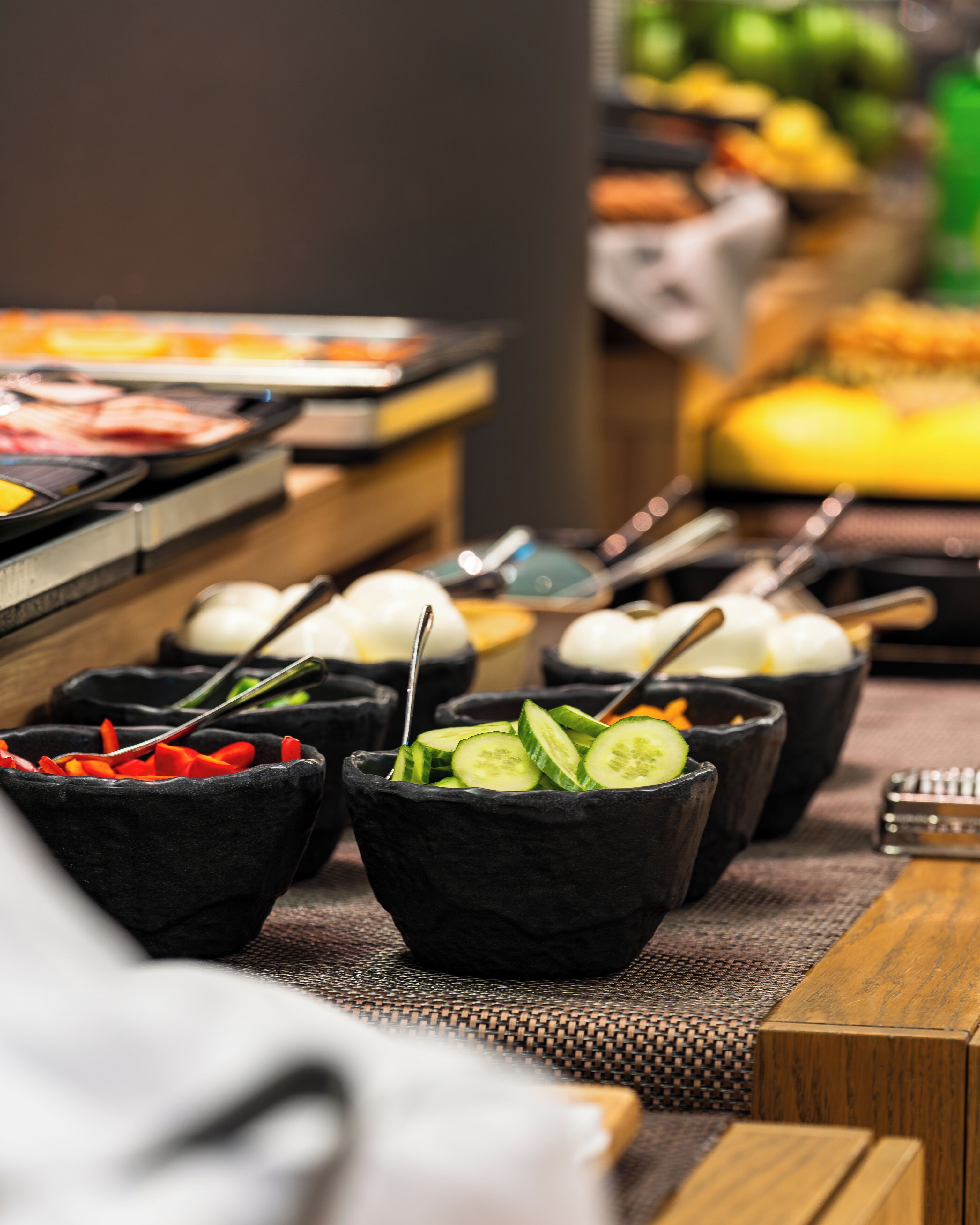 Een buffet met diverse gerechten, waaronder plakjes komkommer en rode paprika in zwarte kommen, en een selectie aan vleeswaren en kazen op het aanrecht.