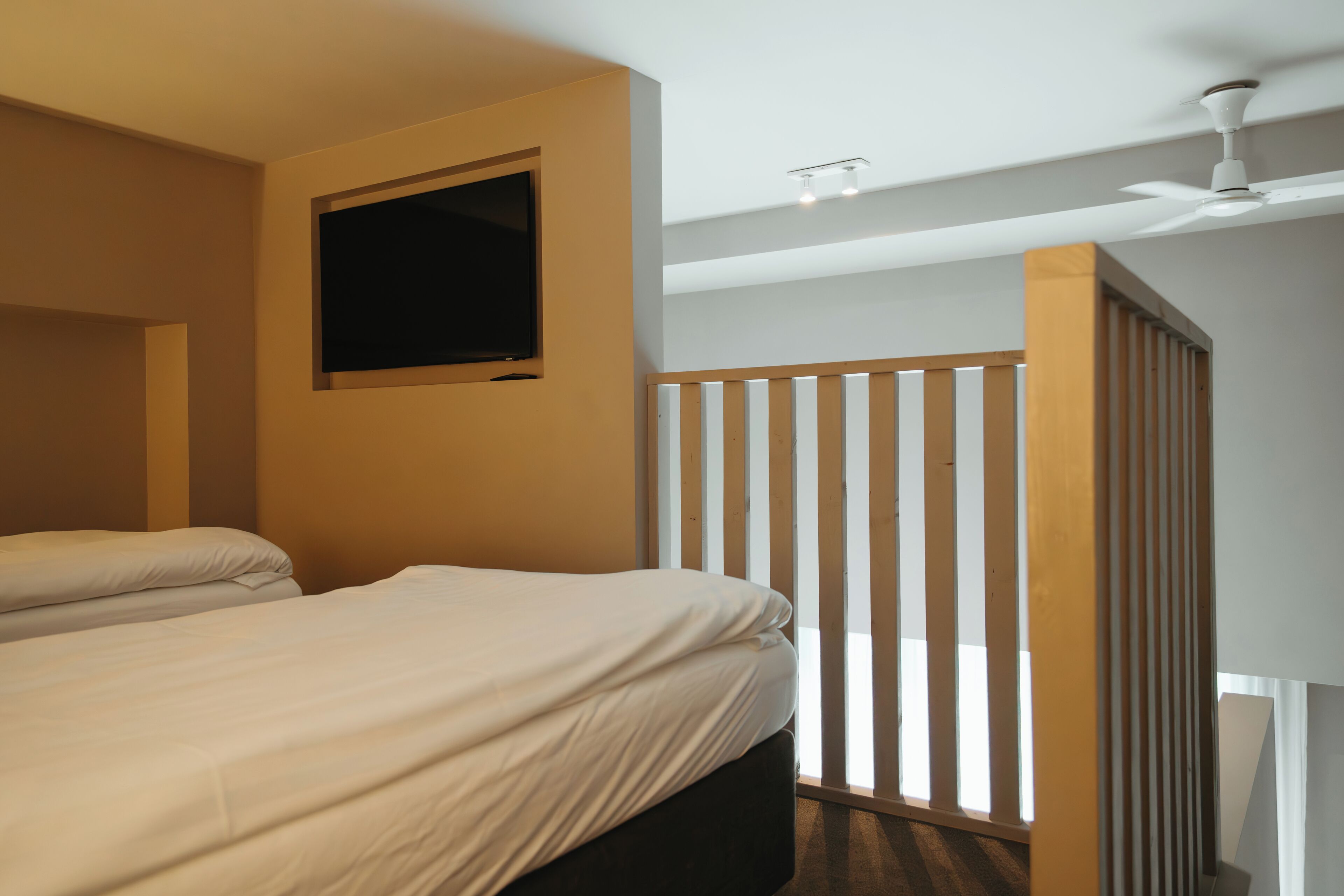 A hotel room with two beds, a ceiling fan, a TV mounted on the wall, and a wooden partition.