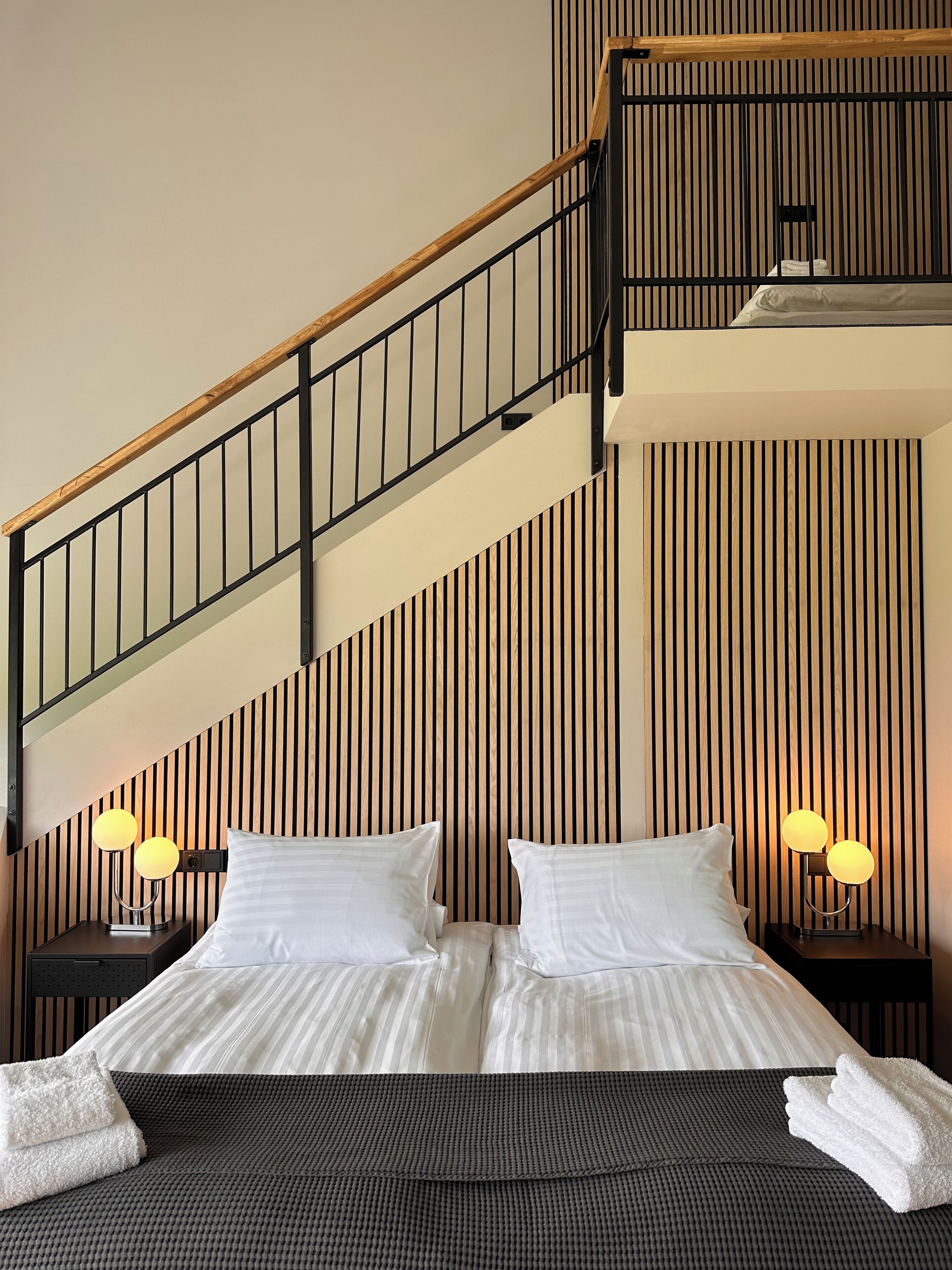 A hotel room with a bed, two bedside lamps, a wooden staircase, and a striped wall.