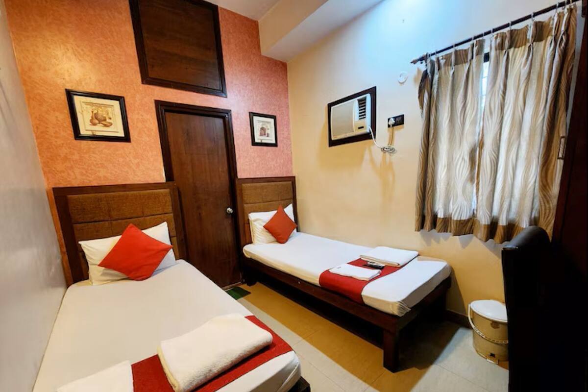Quarto de hotel com duas camas, cabeceira de madeira, pequena janela com cortinas, ar condicionado fixado na parede e uma cadeira de madeira.