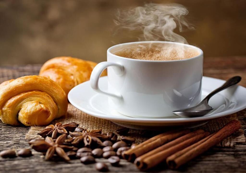 A cup of hot beverage with steam, a croissant, cinnamon sticks, and coffee beans on a wooden surface.