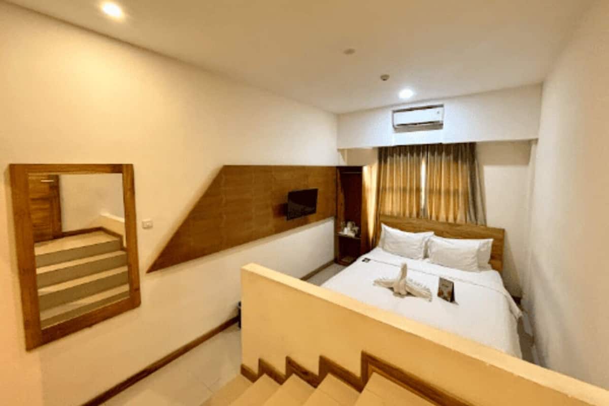 Une chambre d’hôtel avec un lit, une tête de lit en bois, un miroir, un escalier et une fenêtre avec des rideaux.