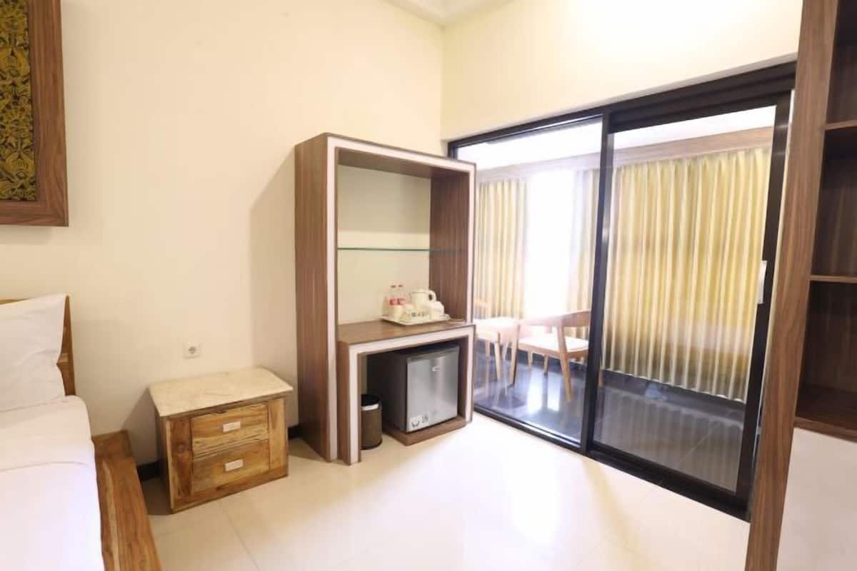 Une chambre d’hôtel comprenant un lit, une petite table de chevet en bois, un bureau avec un plateau en verre, un mini-réfrigérateur et une porte-fenêtre coulissante donnant sur un balcon.