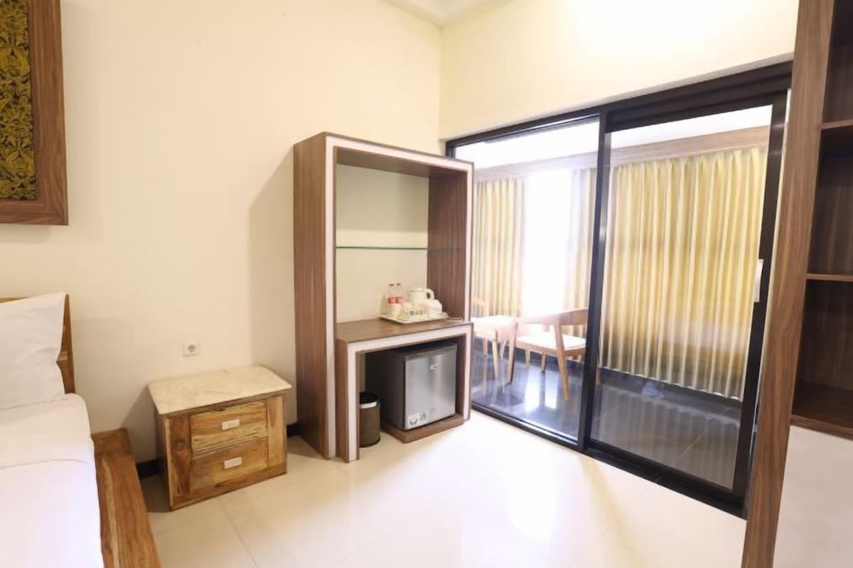 Quarto de hotel com cama, mesa de cabeceira de madeira, escrivaninha com tampo de vidro, mini-geladeira e porta de vidro corria para uma varanda.