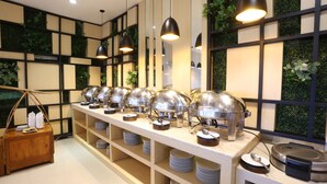 A buffet setup with multiple serving stations, shelves with stacked plates, and a wooden side table with a basket. - Luminor Hotel Banyuwangi Yos Sudarso by WH (Banyuwangi)