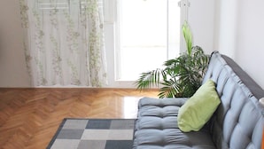 Quarto com sofá cinzento, tapete estampado, planta em vaso e porta que dá para o exterior.