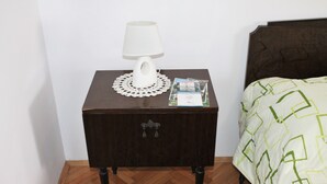 Uma pequena mesa de cabeceira de madeira com uma luminária de mesa branca, um rendado e um livro ao lado de uma cama com colcha verde estampada em padrão geométrico.