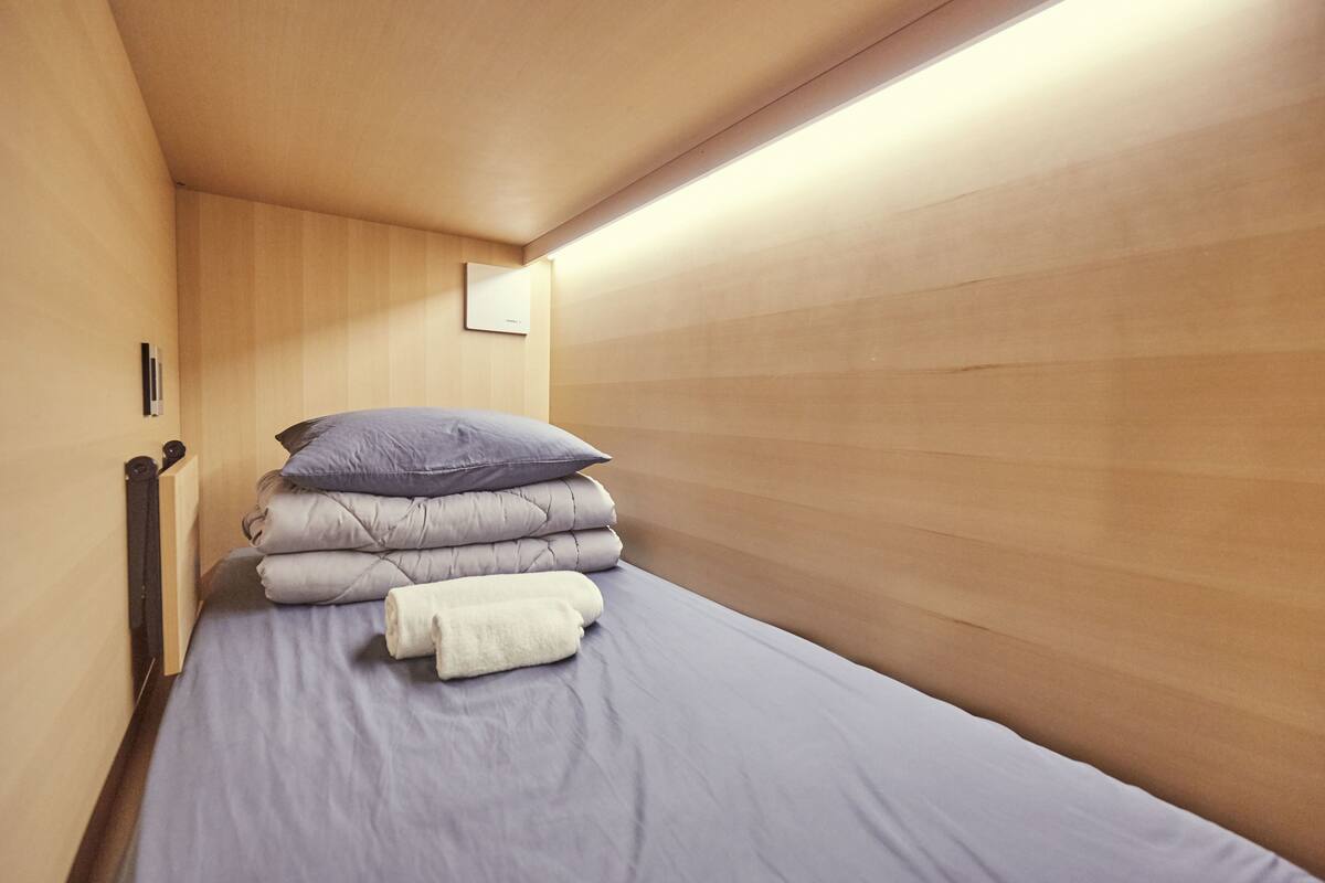 Una cama individual con un colchón gris, una pila de almohadas y una toalla enrollada.