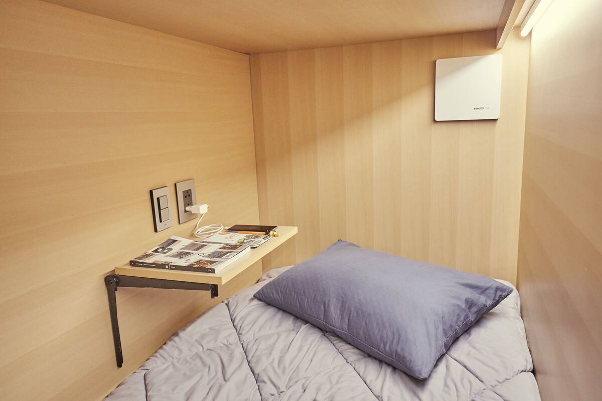 Una habitación pequeña y funcional con cama, mesita de noche con lámpara y aire acondicionado instalado en la pared.