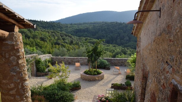 Une cour rustique aux murs de pierre, un petit jardin et une vue sur une chaîne de montagnes boisées.