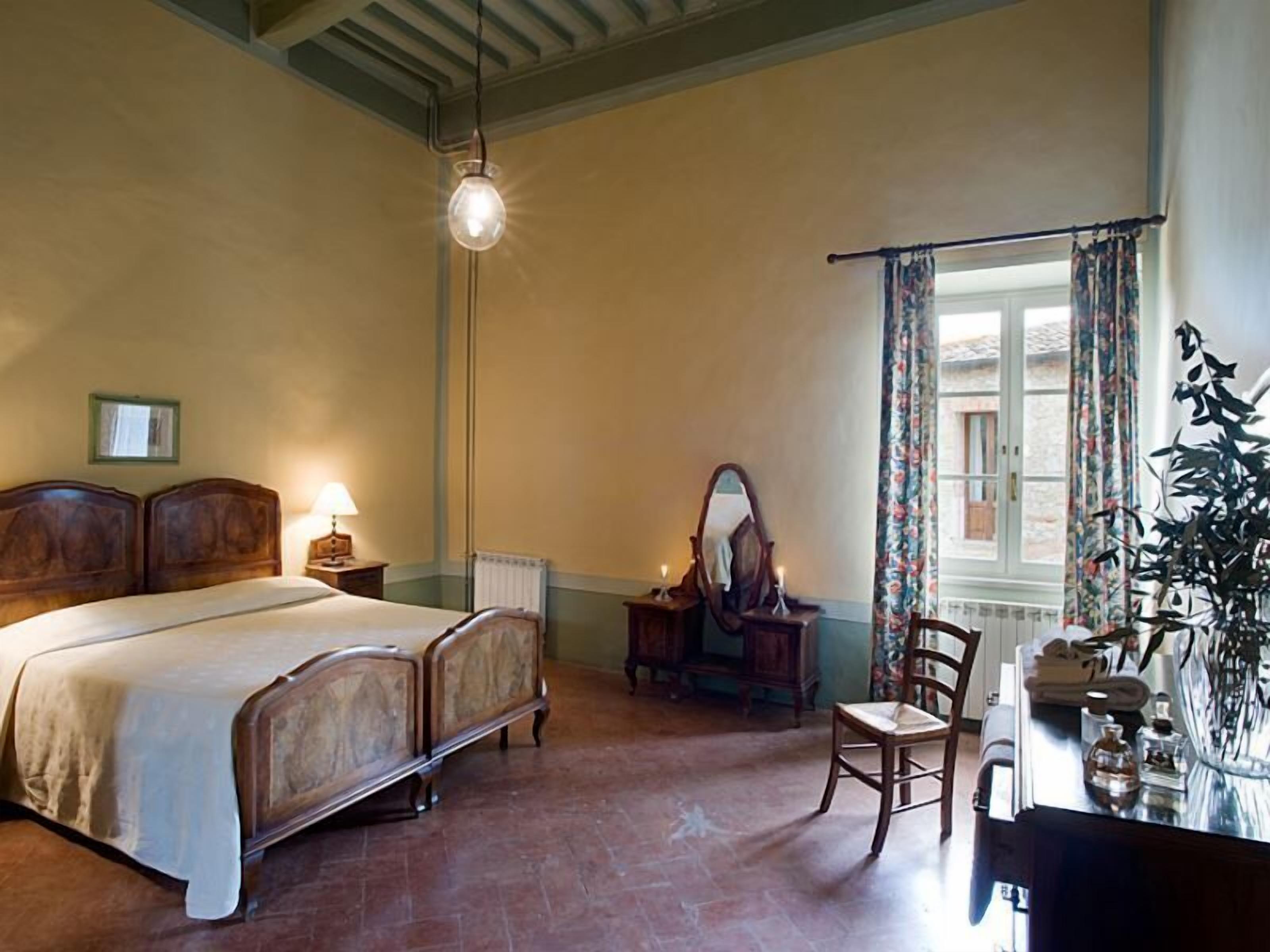 Une chambre à coucher de style traditionnel, comprenant un lit, une table de chevet, une lampe, un miroir, une chaise et une fenêtre avec des rideaux.