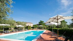 Une piscine entourée de verdure et de bâtiments en pierre, avec des chaises longues et des parasols.