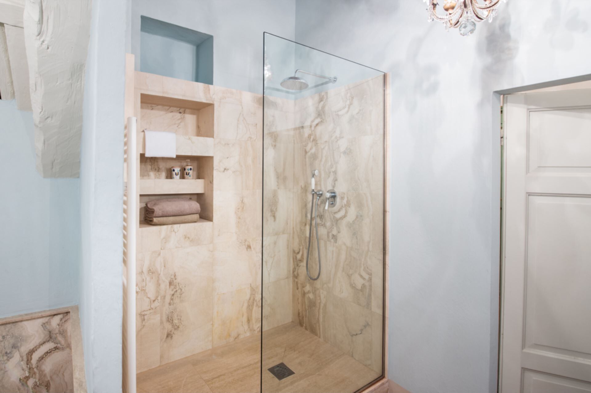 Une douche fermée par des parois de verre, avec des murs en marbre, une étagère pour les serviettes et une porte sur le côté droit.