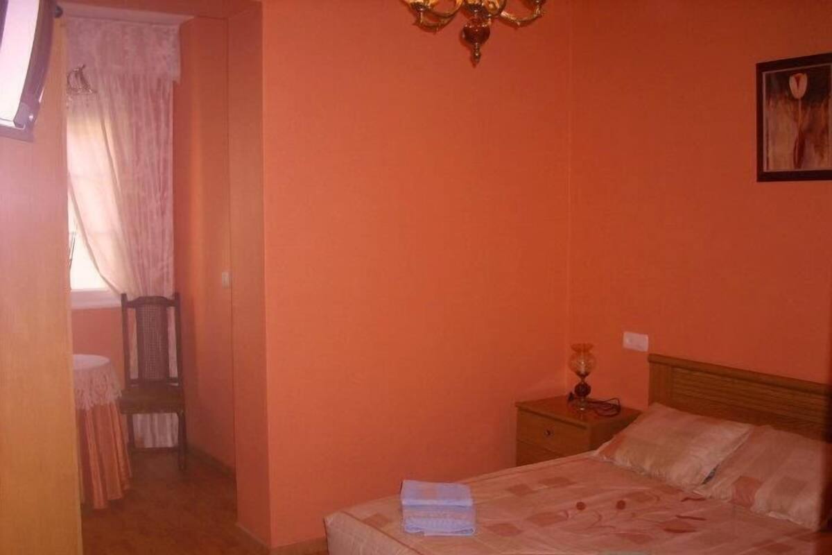 Un dormitorio con cama, mesita de noche, silla y ventana con cortinas.