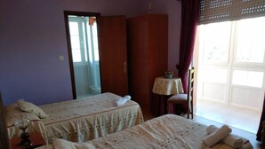 A hotel room with two beds, a wardrobe, a small table with a floral tablecloth, and a chair. - Hostal Cachiño (Carnota)