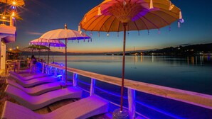 Terraza frente al agua con sillones y sombrillas, con vista a una superficie acuática tranquila.