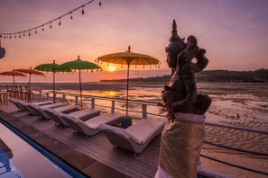Terraza con sillones de descanso y sombrillas que da vista a un atardecer sobre el agua, con una estatua al frente.