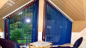 A room with a table and two chairs, large windows with blue blinds, and a view of greenery outside. - Ferienhaus & Nurdachhaus Rothaargebirge (Bad Berleburg)