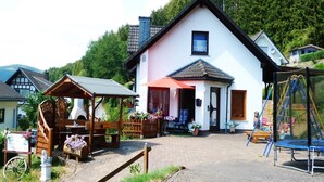 A white building with a black roof, a wooden structure with a roof, and a playground with a swing set and a trampoline. - Ferienhaus & Nurdachhaus Rothaargebirge (Bad Berleburg)
