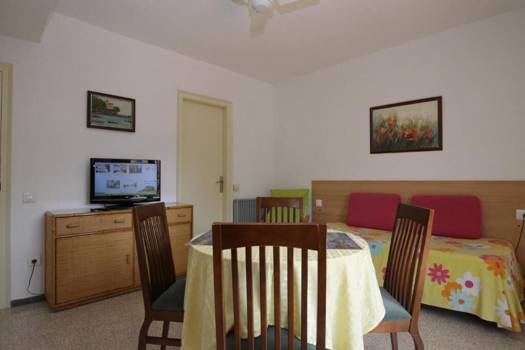 Une pièce comprenant un lit, un téléviseur posé sur un meuble en bois, une table à manger avec des chaises, et deux tableaux accrochés aux murs.
