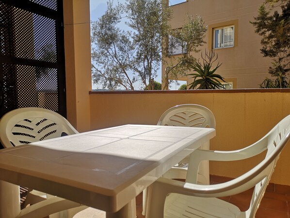 A balcony with a table and chairs, a view of trees and a building, and a tiled floor. - Es Pujols: Apartment in a residence (Es Pujols)