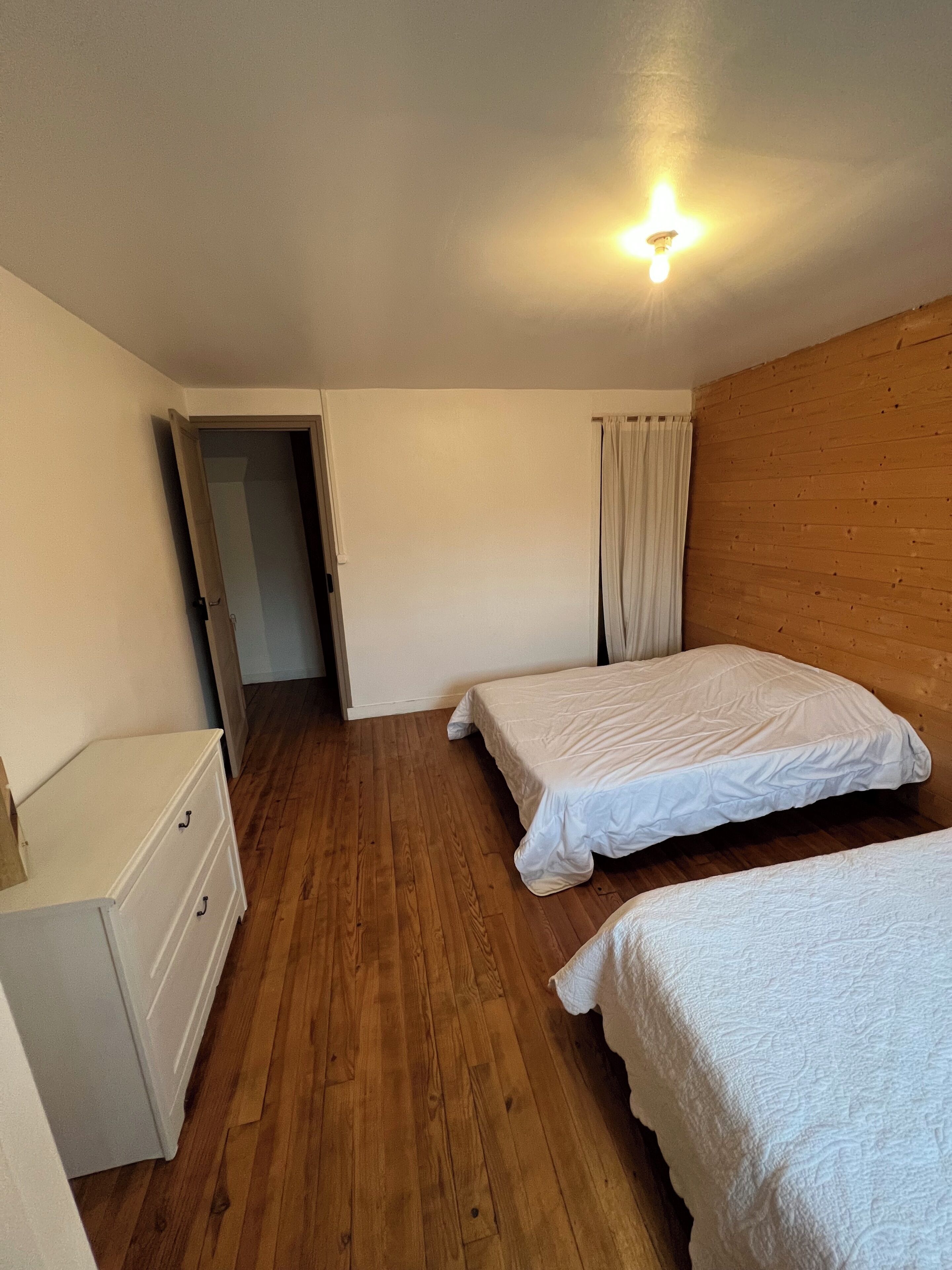 Una habitación con dos camas, piso de madera, un aparador blanco y una pared de madera.