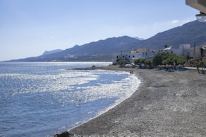 Eine Küstenlandschaft mit Kieselstrand, klarem blauen Meer und Gebäuden vor einer Bergkulisse.