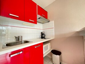 A kitchen with red cabinets, a white countertop, a stainless steel sink, a coffee maker, and a trash bin.