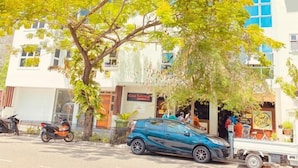 A multi-story building with a white facade and blue windows. There is a tree in front of the building, and a blue car parked on the street. - Casa Retreat (Hulhumalé)