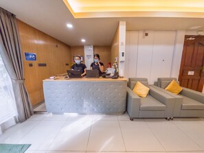 A reception area with a counter, two staff members, a sofa with yellow cushions, and a wooden wall with a clock. - Casa Retreat (Hulhumalé)