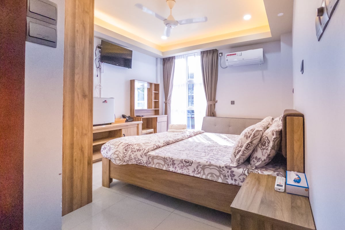 A modern bedroom with a bed, bedside table, and a desk with a chair. There is a TV mounted on the wall and an air conditioning unit.
