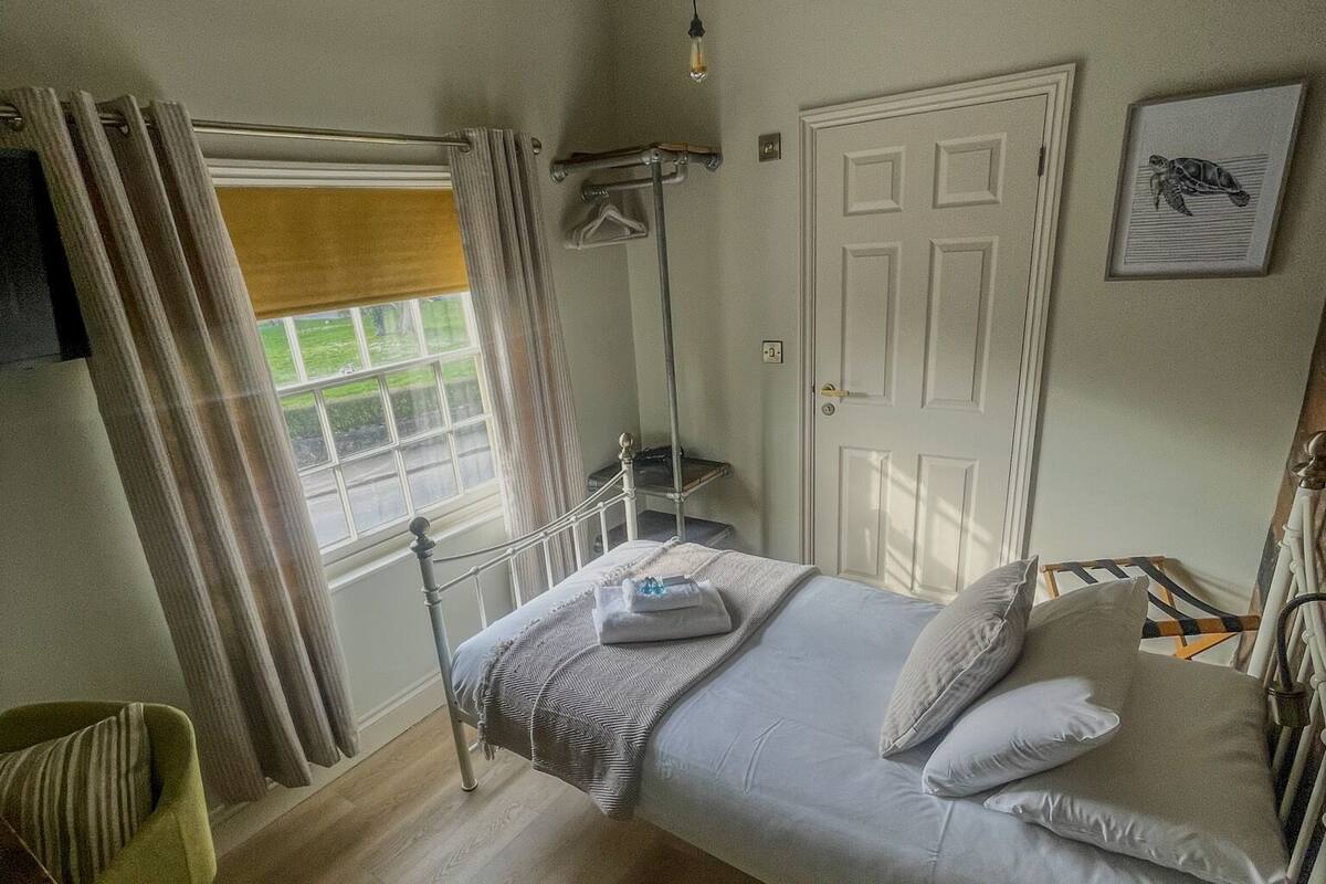 A single bed with pillows and a blanket, a bedside table, a chair, a standing fan, a window with curtains, and a door.