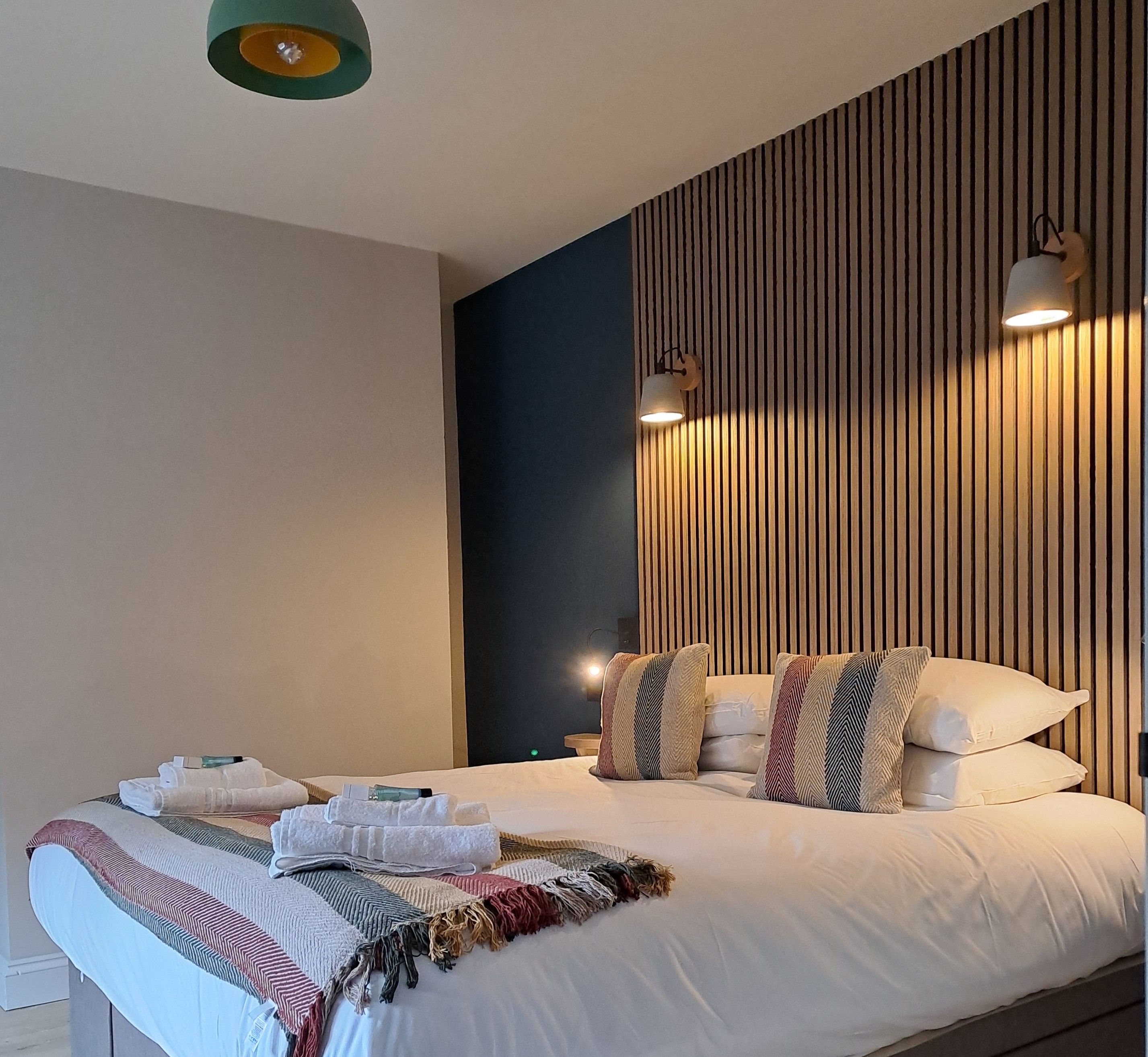 Habitación de hotel con una cama grande, un cabecero de listones de madera y dos lámparas de pared.